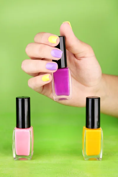 Female hand with stylish colorful nails holding bottle with nail polish, on green background — Stock Photo, Image