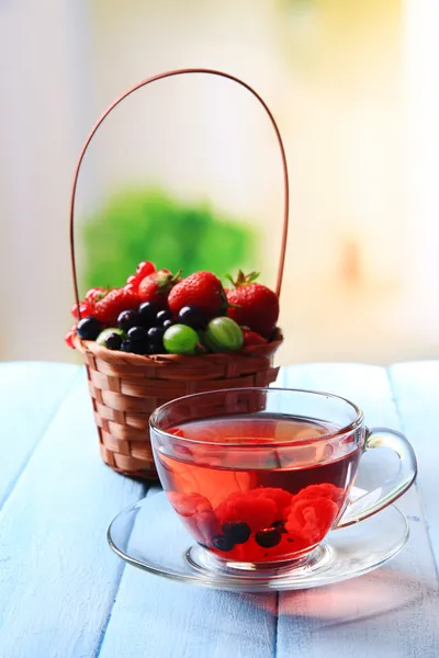 Forest berries in wicker basket and glass cup with red fruit tea, on wooden table, on bright background — Stock Photo, Image