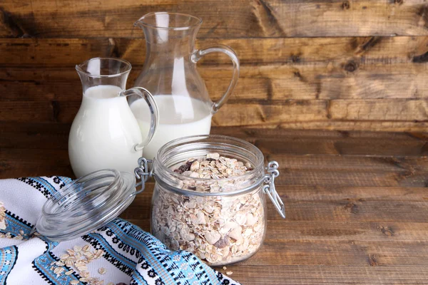 Granola fatta in casa in vaso di vetro, brocche con latte e mela fresca, su fondo di legno a colori — Foto Stock