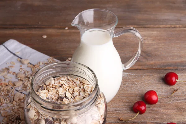 Granola fatta in casa in vaso di vetro, ciliegie fresche e brocca con latte su sfondo di legno di colore — Foto Stock