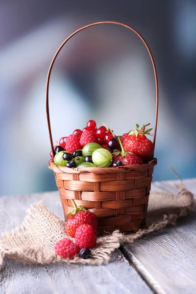 Forest berries in wicker basket, on wooden table, on bright background — Stock Photo, Image