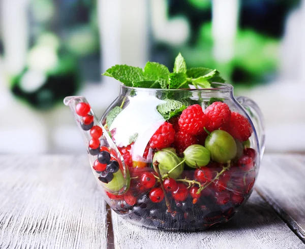 Forest berries and mint leaves in glass teapot, on wooden table, on bright background — Stock Photo, Image