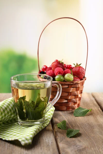 Bessen in rieten mand en glas cup met mint thee, op houten tafel, op lichte achtergrond — Stockfoto