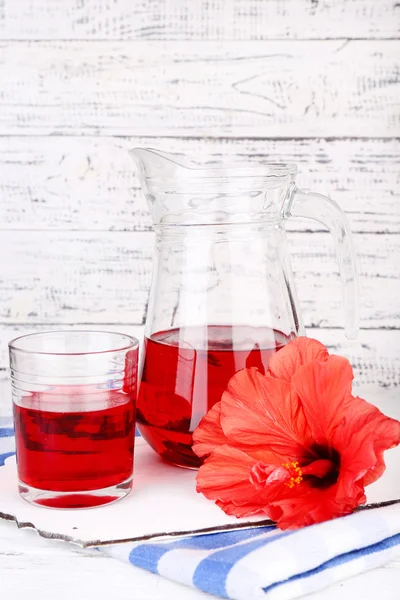Cold hibiscus tea in glass jug with hibiscus flower on wooden background — Stock Photo, Image