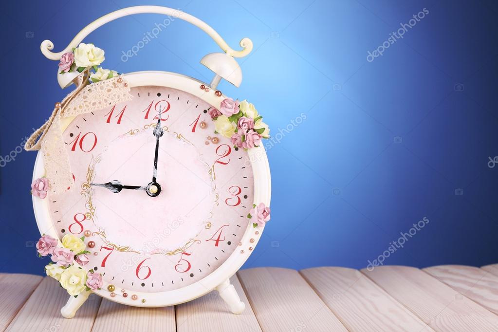 Beautiful vintage alarm clock with flowers on wooden table, on blue background