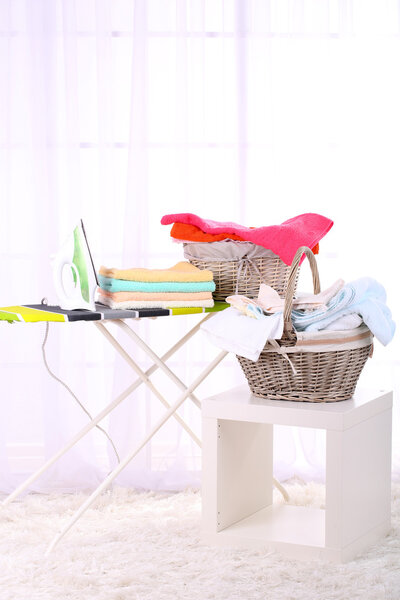 Basket with laundry and ironing board on light home interior background