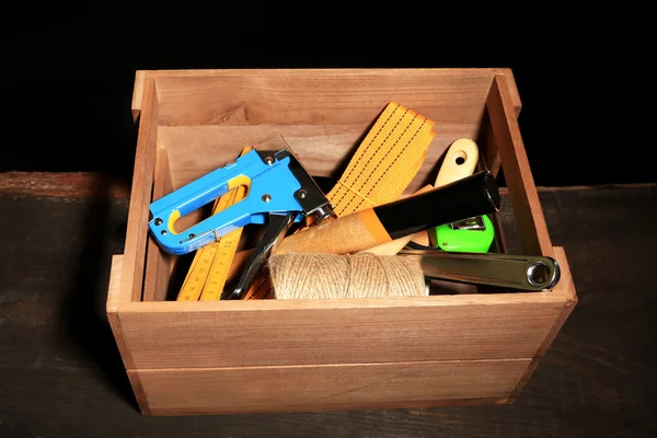 Wooden box with different tools, on wooden table, on dark background — Stock Photo, Image