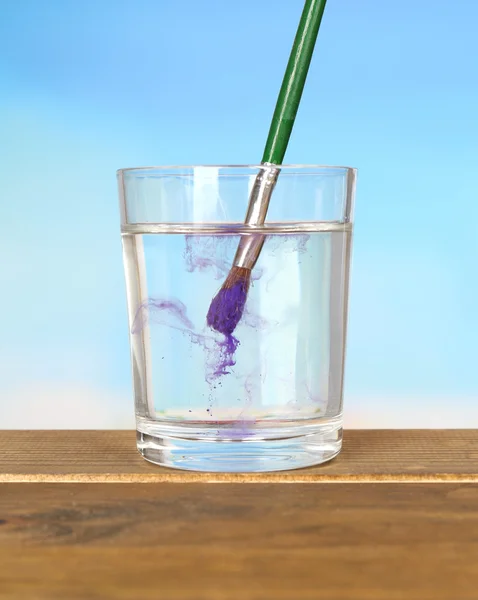 Cepillo con pintura de color en vaso de agua, sobre mesa de madera, sobre fondo azul —  Fotos de Stock