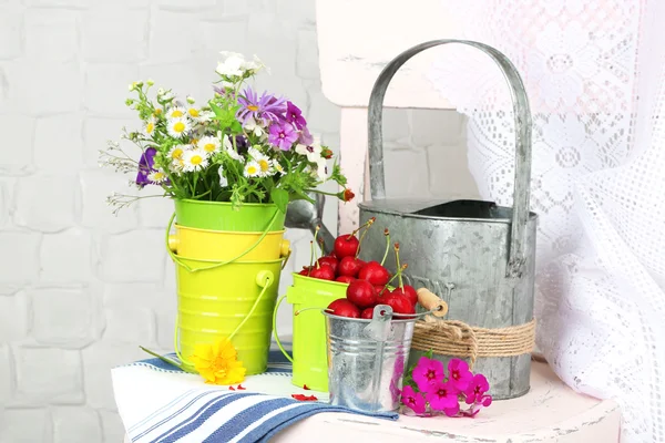 Bukett av färgglada blommor och färska körsbär i dekorativa hinkar, på stol, på ljus vägg bakgrund — Stockfoto