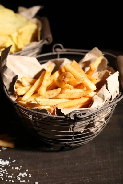 Goda pommes frites i metall korg och potatischips på träbord med mörka ljus — Stockfoto