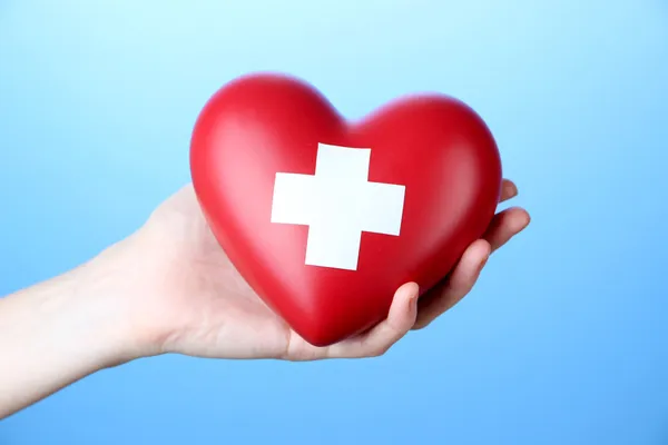 Corazón rojo con signo de cruz en la mano femenina, primer plano, sobre fondo de color — Foto de Stock