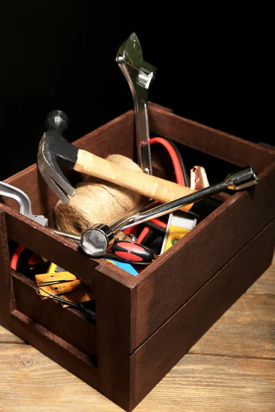 Wooden box with different tools, on wooden table, on dark background — Stock Photo, Image