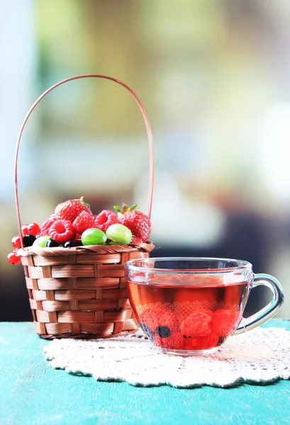 Forest berries in wicker basket and glass cup with red fruit tea, on wooden table, on bright background — Stock Photo, Image