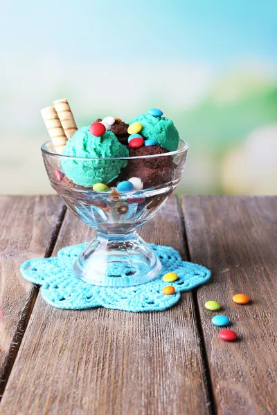 Gelato al cioccolato con caramelle multicolore e rotoli di wafer in ciotola di vetro, su tavolo di legno a colori, su sfondo brillante — Foto Stock