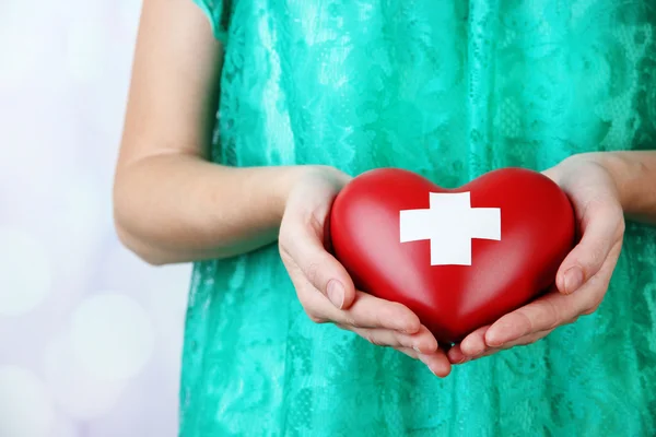 Red heart with cross sign in female hand, close-up, on light background — Stock Photo, Image