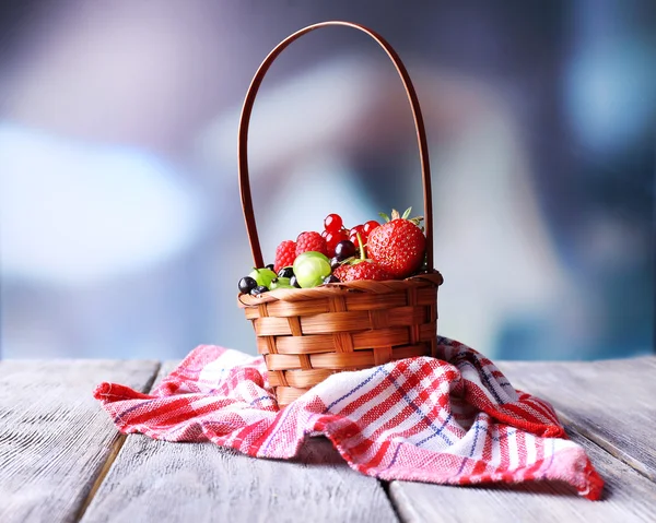 Forest berries in wicker basket, on wooden table, on bright background — Stock Photo, Image