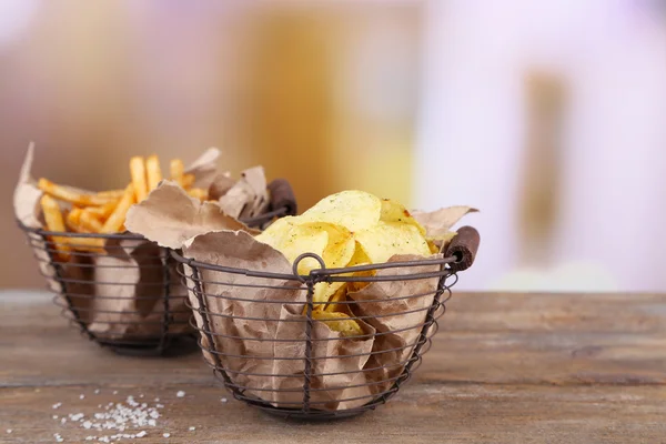 Tasty potato chips and french fries in metal baskets on wooden table, on light background — Stock Photo, Image
