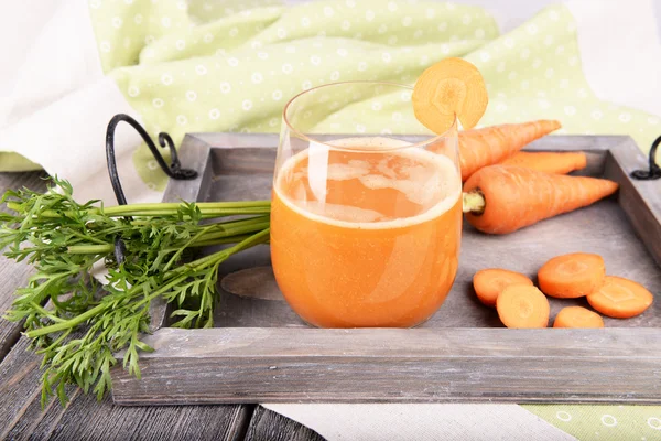 Стакан морковного сока и свежая морковь на деревянном подносе на столе рядом — стоковое фото