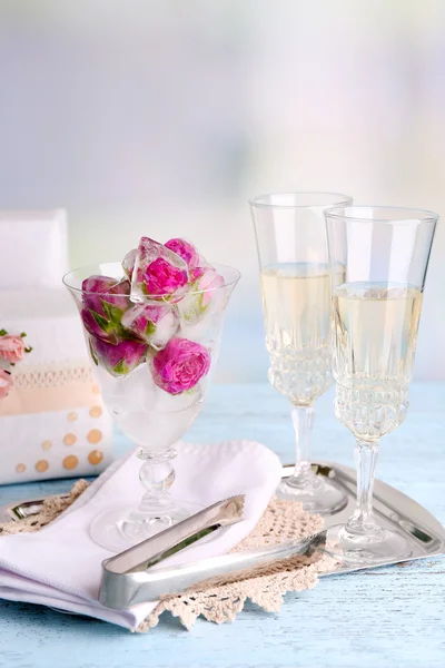 Ледяные кубики с цветами роз в стеклянной чаше и два бокала с шампанским на деревянном столе, на светлом фоне — стоковое фото