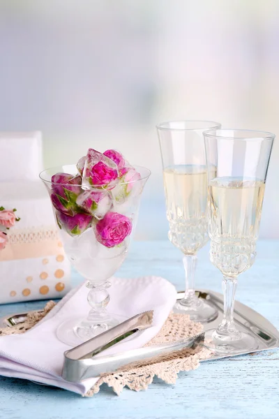 Ice cubes with rose flowers in glass bowl and two glasses with champagne on wooden table, on light background — Stock Photo, Image