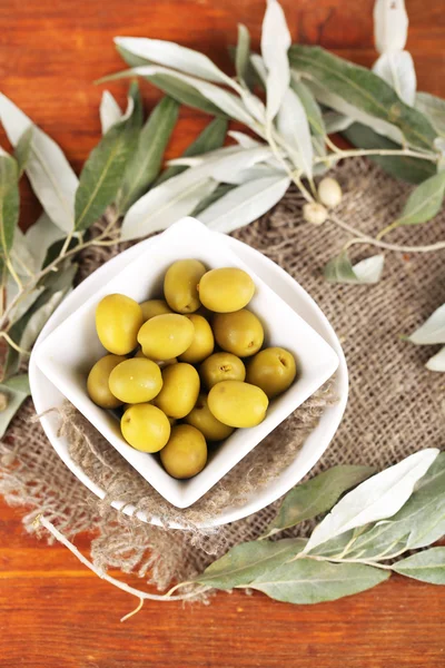 Oliven i skål med gren på sæk på træbord - Stock-foto