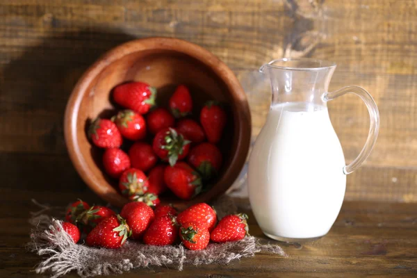 Rijpe zoete aardbeien in houten kom en kruik met melk op een houten achtergrond kleur — Stockfoto