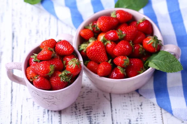 Rijpe zoete aardbeien in pot en mok op een houten achtergrond kleur — Stockfoto