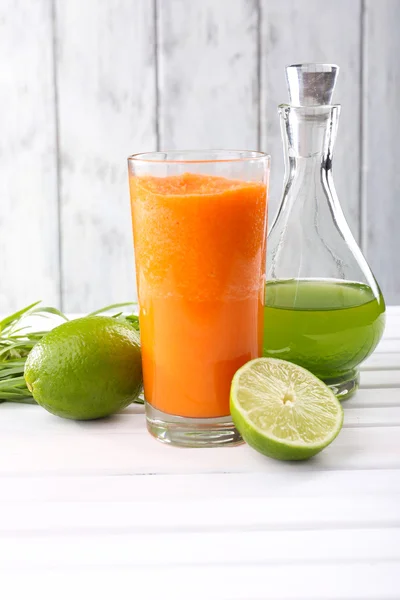 Copo de suco de cenoura fresca, decantador de suco de limão, limão, tufo de grama na mesa em frente à parede de madeira — Fotografia de Stock