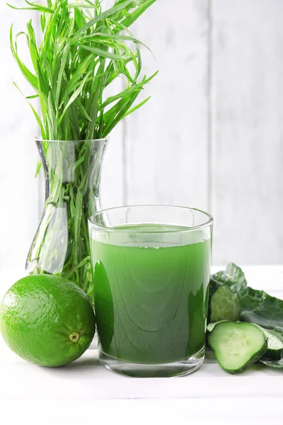 Glass of fresh lime juice, lime, slices of cucumbers and vase of grass on the table in front of wooden wall — Stock Photo, Image
