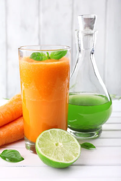 Glass of fresh carrot juice, decanter of lime juice, lime,  carrot, tuft of grass on the table in front of wooden wall — Stock Photo, Image