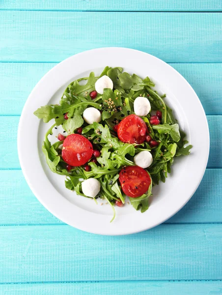 Grüner Salat mit Rucola, Tomaten, Käse-Mozzarella-Bällchen und Sesam auf Teller, auf farbigem Holzhintergrund — Stockfoto