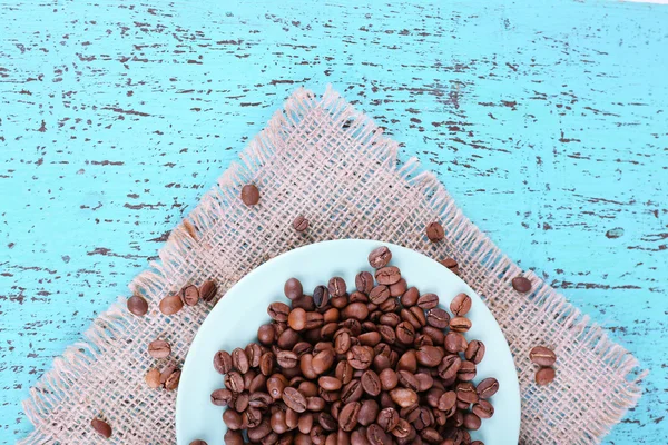 Ziarna kawy w białym talerzu na serwetce workowej na niebieskim drewnianym tle — Zdjęcie stockowe