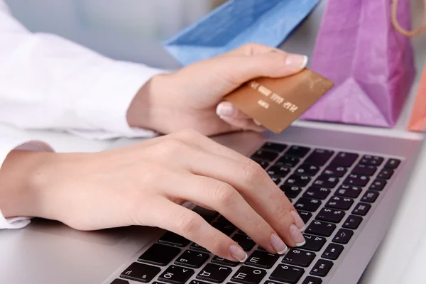 Mãos femininas segurando cartão de crédito com laptop e sacos de papel na mesa em fundo brilhante — Fotografia de Stock