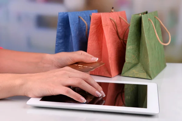 Mãos femininas segurando cartão de crédito com tablet de computador e sacos de papel na mesa em fundo brilhante — Fotografia de Stock