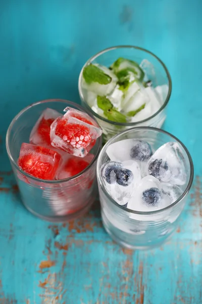 Ice cubes with mint leaves, raspberry and blueberry in glasses, on color wooden background — Stock Photo, Image