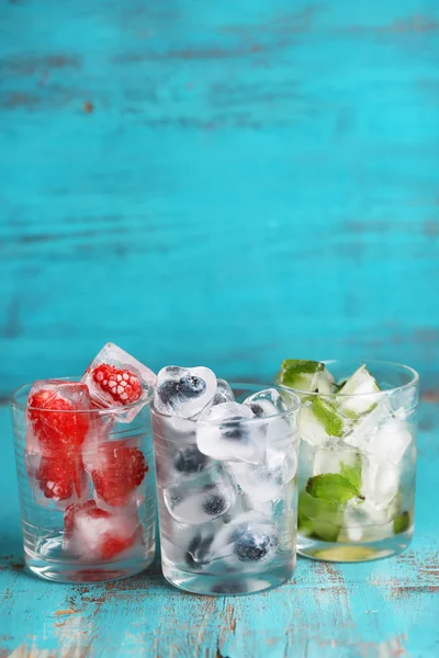Cubi di ghiaccio con foglie di menta, lampone e mirtillo in bicchieri, su sfondo di legno a colori — Foto Stock