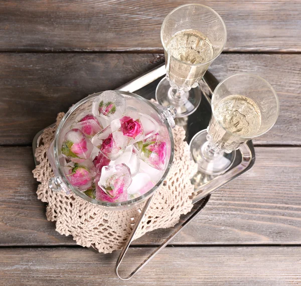 Ice cubes with rose flowers in glass bucket and two glasses with champagne on wooden table background — Stock Photo, Image