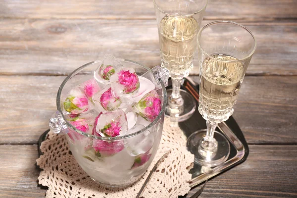 Ijsblokjes met roze bloemen in glas emmer en twee glazen met champagne op houten tafel achtergrond — Stockfoto