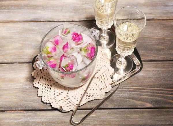 Cubos de gelo com flores de rosa em balde de vidro e dois copos com champanhe em fundo de mesa de madeira — Fotografia de Stock