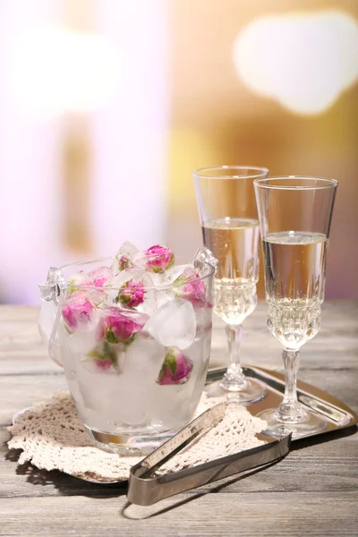 Cubetti di ghiaccio con fiori di rosa in secchio di vetro e due bicchieri con champagne su tavolo di legno, su sfondo luminoso — Foto Stock