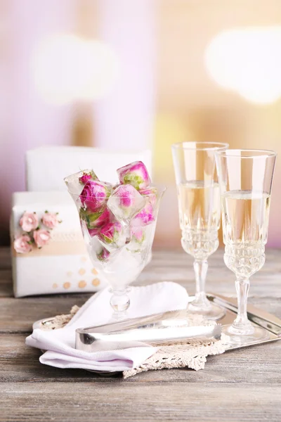 Ледяные кубики с цветами роз в стеклянной чаше и два бокала с шампанским на деревянном столе, на ярком фоне — стоковое фото