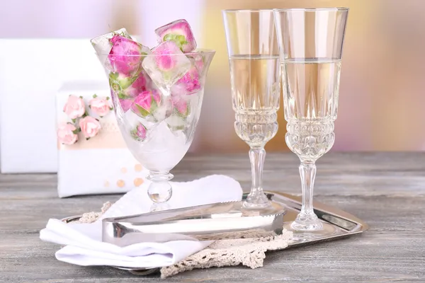 Ice cubes with rose flowers in glass bowl and two glasses with champagne on wooden table, on bright background — Stock Photo, Image