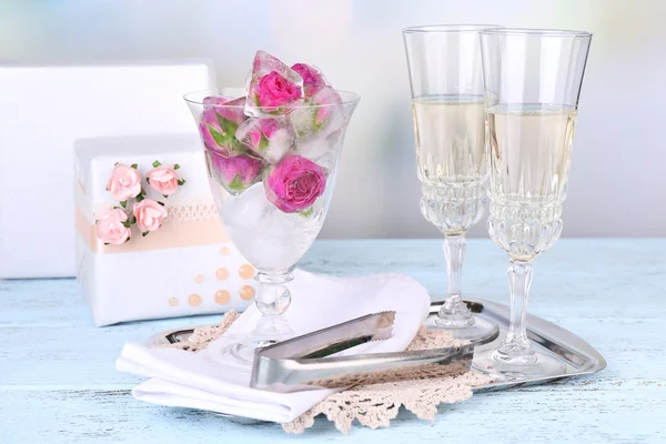 Παγάκια με τα ροδαλά λουλούδια σε γυάλινο μπολ και δύο ποτήρια με σαμπάνια στο ξύλινο τραπέζι, επάνω ελαφρύς υπόβαθρο — Φωτογραφία Αρχείου