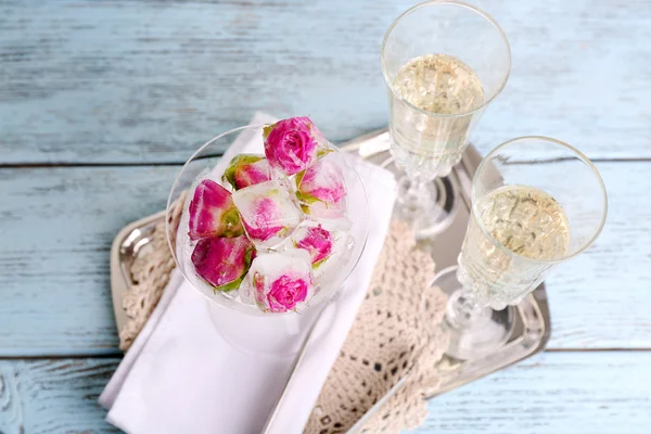 Ice cubes with rose flowers in glass bowl and two glasses with champagne on wooden table, on light background — Stock Photo, Image