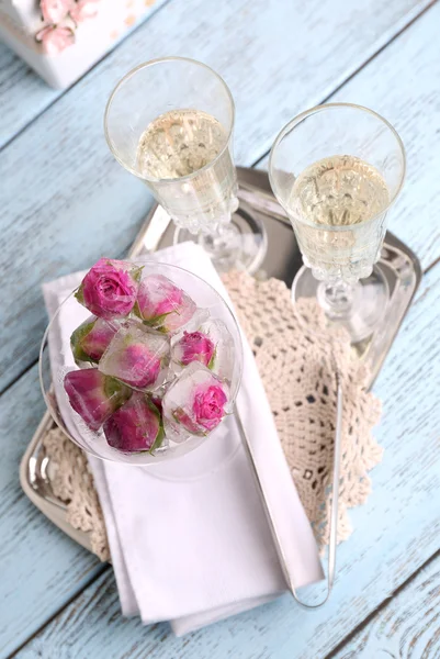 Ice cubes with rose flowers in glass bowl and two glasses with champagne on wooden table, on light background — Stock Photo, Image