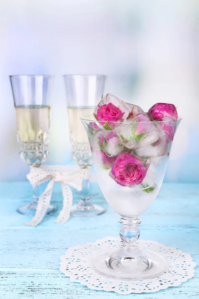 Cubetti di ghiaccio con fiori di rosa in ciotola di vetro e due bicchieri con champagne sul tavolo di legno, su sfondo chiaro — Foto Stock