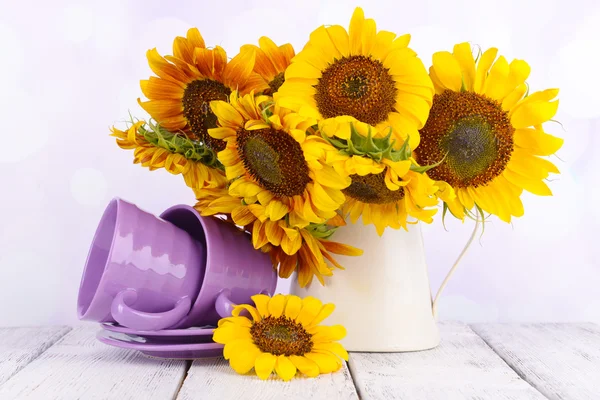 Beautiful sunflowers in pitcher with cups on table on bright background — Stock Photo, Image