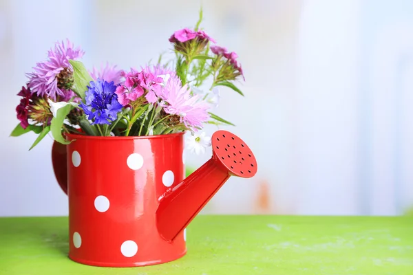 Bright wildflowers in decorative watering can, on wooden table, on bright background — Stock Photo, Image