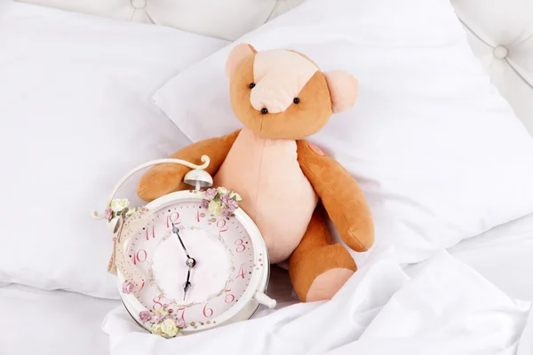 Metal clock and toy bear on pillows on a big white bed — Stock Photo, Image