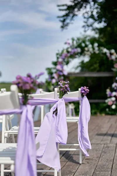 White wedding chairs with fresh flowers and purple cloth on each side of archway outdoods, copy space.  Empty wooden chairs prepared for wedding ceremony